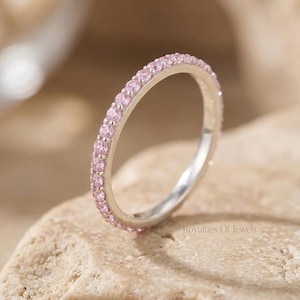 Può includere: Un anello d'argento con una fascia di piccole pietre preziose rosa. L'anello è appoggiato su una superficie chiara e strutturata. Un gioiello delicato.