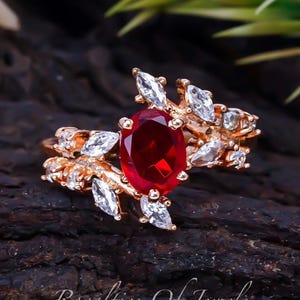 May include: A gold ring with a large red oval gemstone surrounded by smaller clear gemstones. The ring features a delicate leaf design.