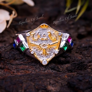 May include: A silver and gold ring with a shield design featuring a stylized bird and a triangle with an eye inside. The shield is surrounded by small clear stones and the band is decorated with four square stones in red, green, and blue.