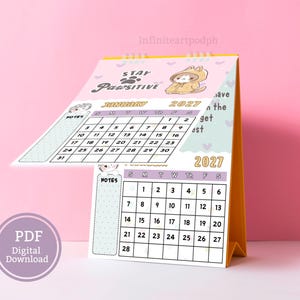 May include: A 2027 desk calendar with a spiral binding and a yellow stand. The pages feature a light pink background with a cartoon cat in a hoodie, the text Stay Pawsitive, and monthly grids for January and February with a notes section on the left.
