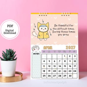May include: A spiral-bound wall calendar for April 2027 featuring a cartoon cat in a yellow raincoat under rain clouds. The text reads: Be thankful for the difficult times. During those times you grow. It includes a grid for dates and a notes section.