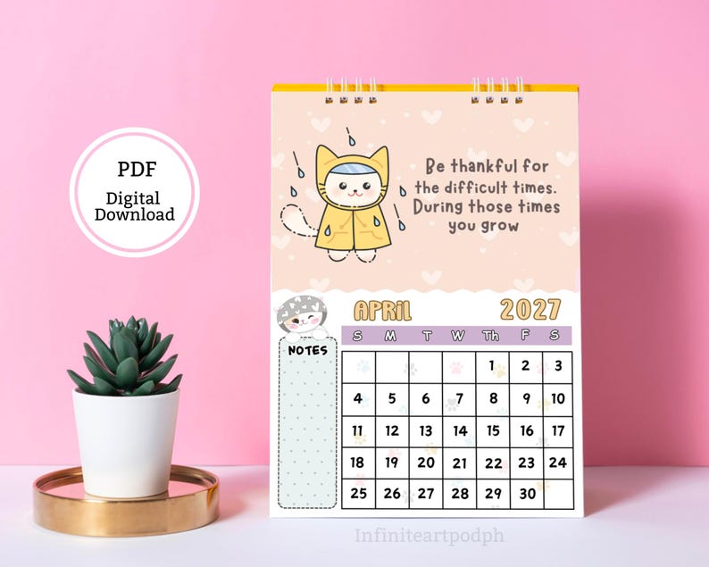 May include: A spiral-bound wall calendar for April 2027 featuring a cartoon cat in a yellow raincoat under rain clouds. The text reads: Be thankful for the difficult times. During those times you grow. It includes a grid for dates and a notes section.