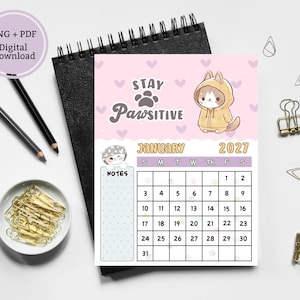 May include: A January 2027 calendar page featuring a light pink background with purple hearts, a cartoon cat in a yellow hoodie, and the text Stay Pawsitive. The calendar includes a grid for dates, a notes section, and a small cat illustration.