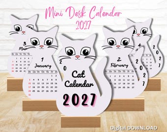 May include: A set of cat-shaped mini desk calendars for 2027, each featuring a white cat illustration with large eyes and a wooden base. The calendars display months including January, February, and March, with the text Cat Calendar 2027 visible.