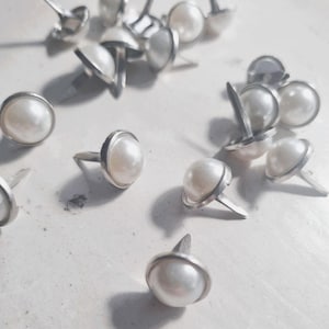 Puede incluir: Una colección de tachuelas plateadas para tapicería con cabezas de perlas de imitación. Las tachuelas tienen un centro de perla redondeado y abovedado, un borde de metal plateado y un vástago largo y puntiagudo.