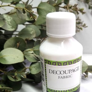 Op de afbeelding: Een witte fles decoupage-stoflijm met een witte dop. Het etiket bevat de tekst "DECOUPAGE FABRIC" in het zwart. Groen gebladerte staat op de achtergrond.