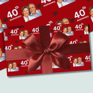 May include: A gift wrapped in red paper with a repeating pattern of a couple's photo and the text "40th Anniversary." A large burgundy satin bow adorns the gift, creating a festive presentation. Small brown heart shapes are scattered across the wrapping paper.