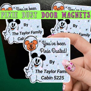 May include: Square magnets with a light blue glitter background featuring a white ghost character holding an orange pumpkin balloon with mouse ears. The text reads You've been Pixie Dusted! By The Taylor Family Cabin 5225 with small black star accents.