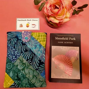 May include: Handmade book sleeve crafted from colourful patterned fabric. The sleeve is folded and laid flat on a pink surface. A book titled "Mansfield Park" by Jane Austen is also pictured.