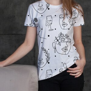 May include: White t-shirt with a black line drawing pattern of faces, flowers, and wine glasses. The pattern is repeated throughout the shirt.