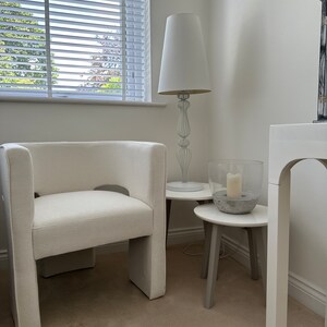 May include: A white upholstered armchair with a unique cutout design sits in a white room with a white lamp and two white side tables. The room has white walls and a white floor.