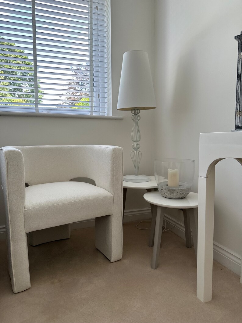 May include: A white upholstered armchair with a unique cutout design sits in a white room with a white lamp and two white side tables. The room has white walls and a white floor.