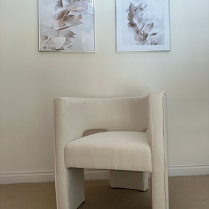 May include: A white upholstered armchair with a unique, modern design. The chair has a high back and a wide seat, and the legs are curved and slightly angled. The chair is sitting on a beige carpet in front of a white wall with two framed prints of botanical illustrations.