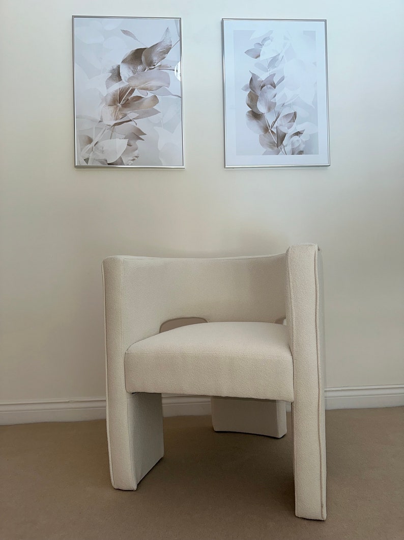 May include: A white upholstered armchair with a unique, modern design. The chair has a high back and a wide seat, and the legs are curved and slightly angled. The chair is sitting on a beige carpet in front of a white wall with two framed prints of botanical illustrations.