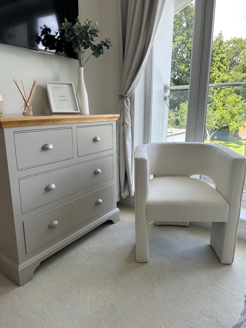 May include: A gray dresser with four drawers and a white chair with a white cushion. The chair is in front of a window with white curtains. The dresser has a sign on top that says "breathe in, exhale" in cursive.