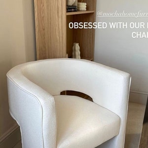 May include: A white upholstered armchair with a unique curved design and a cutout in the back. The chair is sitting in a room with a wooden bookshelf and a beige carpet. The text "OBSESSED WITH OUR NEW CHAIR" is visible in the image.