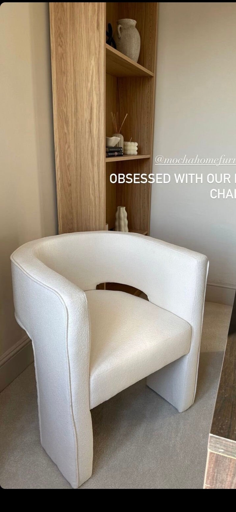May include: A white upholstered armchair with a unique curved design and a cutout in the back. The chair is sitting in a room with a wooden bookshelf and a beige carpet. The text "OBSESSED WITH OUR NEW CHAIR" is visible in the image.