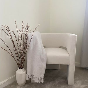 May include: A white armchair with a white throw blanket draped over the back. A vase with pussy willow branches sits on the floor next to the chair.