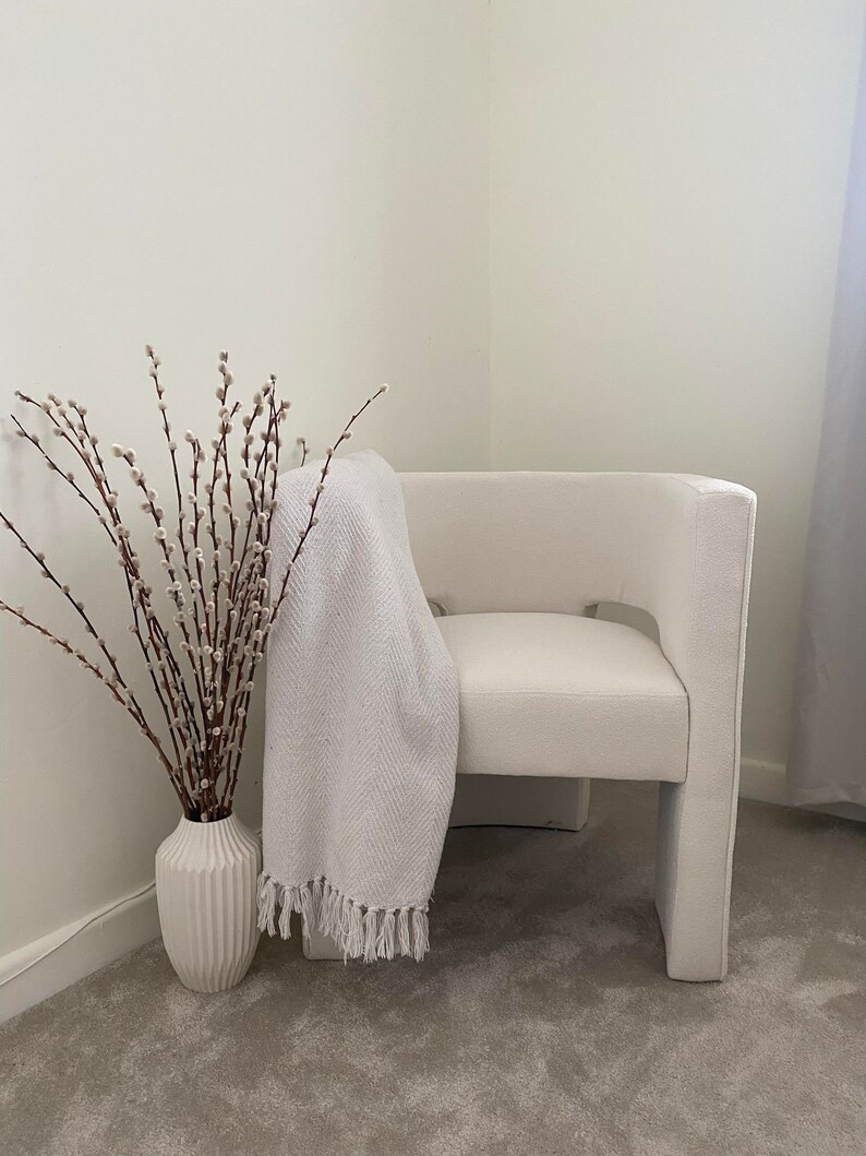 May include: A white armchair with a white throw blanket draped over the back. A vase with pussy willow branches sits on the floor next to the chair.