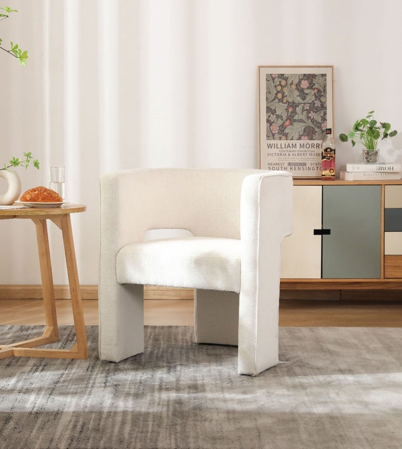 May include: A white upholstered armchair with a curved back and a wide seat. The chair is sitting on a gray rug in a room with white walls and a wooden floor.