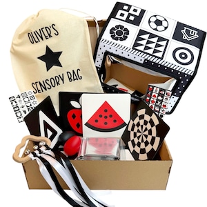 May include: A black and white sensory bag for children, with a black star and the text "Oliver's Sensory Bag" on the front. The bag is filled with various black and white sensory toys, including a wooden rattle, a black and white patterned fabric square, and a set of black and white flash cards.