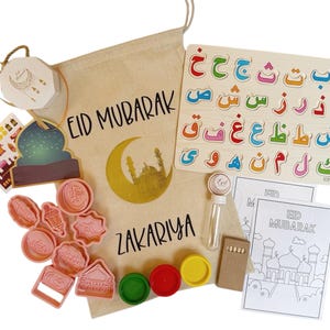 Pode incluir: Uma bolsa de lona bege com o texto "Eid Mubarak" e "Zakariya" impresso nela. A bolsa contém uma variedade de itens, incluindo um quebra-cabeça de alfabeto de madeira com letras árabes, um conjunto de cortadores de biscoitos rosa, uma pequena garrafa de glitter, um livro para colorir com o texto "Eid Mubarak" e um conjunto de massa de modelar.