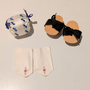 May include: An assortment of doll accessories, including a white satin pouch with blue floral embroidery and a blue ribbon tie. There are also a pair of black leather sandals with wooden soles and a pair of white fabric cuffs with red embroidery.