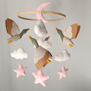May include: A baby mobile featuring felt ducks with brown and gray wings, green heads, and orange beaks. The mobile also includes white clouds, pink stars, and a pink crescent moon, all suspended from a wooden ring.