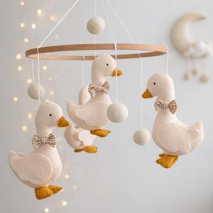 Pode incluir: Móbile para bebé com quatro figuras de pato branco de pelúcia, com bicos e pés amarelos e laços listrados. Os patos penduram-se de um anel de madeira com fio branco e pequenas bolas de feltro branco. Decoração de lua e nuvem no fundo.