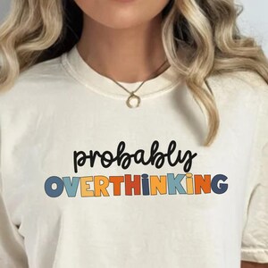 May include: A cream-coloured t-shirt featuring the words "probably OVERTHINKING" in black, blue, orange, and yellow. The shirt has short sleeves and a loose fit, suitable for casual wear.