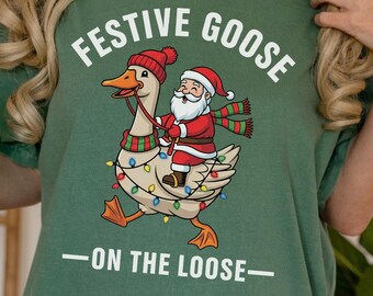 May include: Green t-shirt with the text "Festive Goose" above an illustration of a goose wearing a Santa hat and scarf, with Santa riding on its back. The text "-ON THE LOOSE-" is below the illustration. A green glass jar is held in the left hand.