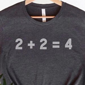 以下が含まれることがあります： 前面に白い文字で「2+2=4」がプリントされた濃いグレーのTシャツです。