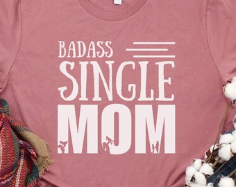 May include: A dusty rose t-shirt with the text "BADASS SINGLE MOM" in white. The shirt is styled with white sneakers, blue jeans, a plaid scarf, and cotton stems. The shirt is folded on a light wood surface.