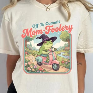 Peut inclure: T-shirt crème avec un graphisme fantaisiste. Le motif représente une grenouille verte portant un chapeau de sorcière, conduisant un scooter rose dans un paysage coloré. Le texte au-dessus du graphisme se lit "Off To Commit Mom-Foolery."