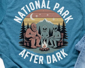 Pode incluir: Camiseta azul-petróleo com um gráfico de Bigfoot e dois alienígenas ao redor de uma fogueira, com o texto "NATIONAL PARK AFTER DARK" em branco. O design inclui uma montanha, árvores, lua e estrelas.