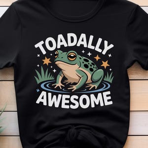 May include: Black t-shirt with a cartoon frog design. The frog is green and tan with black spots, and is in water. The text reads "TOADALLY AWESOME" in white, with stars and grass.