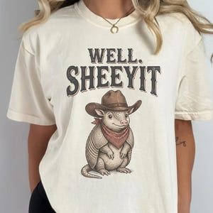 May include: Cream-coloured t-shirt with the text "WELL. SHEEYIT" above an illustration of an armadillo wearing a cowboy hat and a red bandana. The armadillo is standing.