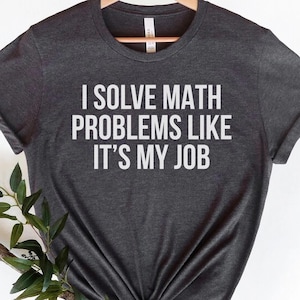 Pode incluir: T-shirt cinzenta escura com o texto branco "I SOLVE MATH PROBLEMS LIKE IT'S MY JOB". A camisa é exibida em um cabide de madeira contra um fundo branco. As mangas são dobradas.