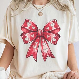 May include: A white t-shirt with a red bow with white hearts printed on it.