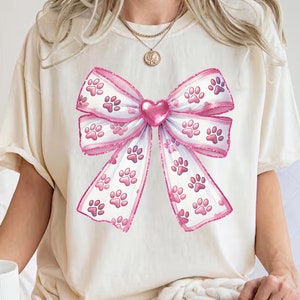 May include: Cream-colored t-shirt featuring a large pink bow design. The bow is adorned with paw prints and a pink heart in the center. The shirt has short sleeves and a relaxed fit, suitable for casual wear.