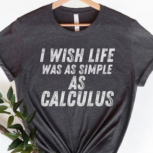 May include: Dark gray t-shirt with the text "I WISH LIFE WAS AS SIMPLE AS CALCULUS" in white, displayed on a wooden hanger. The shirt is folded at the bottom and has rolled-up sleeves. A sprig of greenery is placed on the lower left.