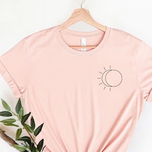 Op de afbeelding: Een lichtroze T-shirt met een simpel zon- en maandesign op de linkerborst. Het shirt hangt aan een hanger en heeft opgerolde mouwen.