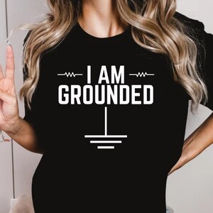 Könnte beinhalten: Ein schwarzes T-Shirt mit dem Text "I AM GROUNDED" in Weiß, mit einem stilisierten Erdsymbol unter dem Text.