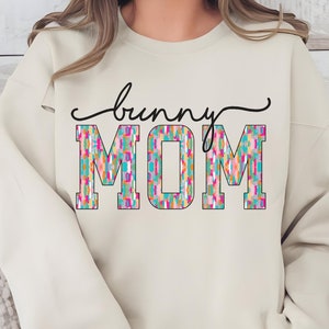 May include: A beige sweatshirt with the text "bunny MOM" in black and colorful patterned letters.
