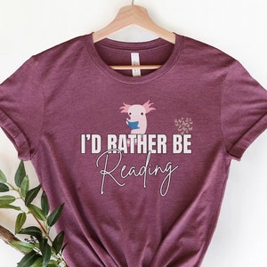 May include: A maroon t-shirt with a white axolotl reading a book and the text "I'd Rather Be Reading".