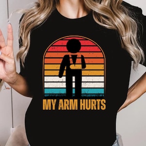 Puede incluir: Camiseta negra con un gráfico de puesta de sol retro de una persona con un brazo roto y el texto "MY ARM HURTS".