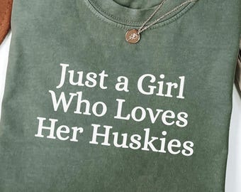 Könnte beinhalten: Ein olivgrünes T-Shirt mit dem weißen Text "Just a Girl Who Loves Her Huskies". Eine silberne Halskette mit einem kleinen Anhänger liegt auf dem Shirt. Das Shirt ist gefaltet und liegt auf einer weißen Oberfläche.