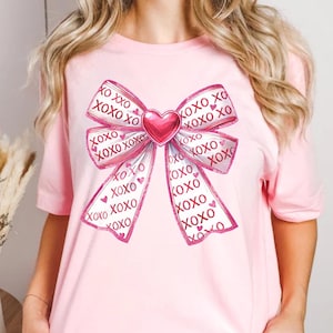 May include: A light pink t-shirt featuring a large bow design. The bow is white with pink trim and the words "XOXO" printed repeatedly. A pink heart is in the center of the bow. The shirt is a short-sleeved crew neck.