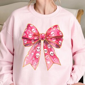May include: Light pink sweatshirt featuring a large pink bow with a skull pattern and gold trim. The bow is the focal point, with a watercolor effect. The sweatshirt is a solid light pink color.