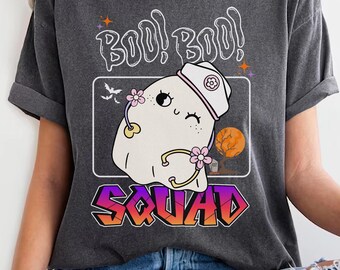 May include: A dark grey t-shirt featuring a cartoon ghost wearing a nurse's hat and holding a stethoscope. The ghost has pink flowers on its head and is enclosed in a white frame. The text "Boo! Boo! Squad" is printed on the shirt.